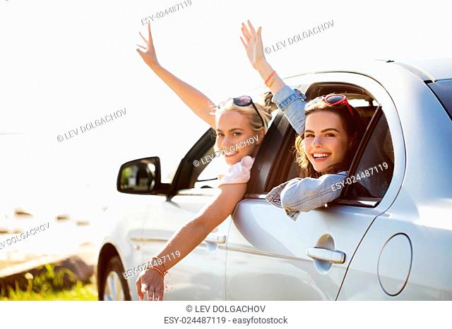 summer vacation, holidays, travel, road trip and people concept - happy teenage girls or young women in car at seaside waving hands