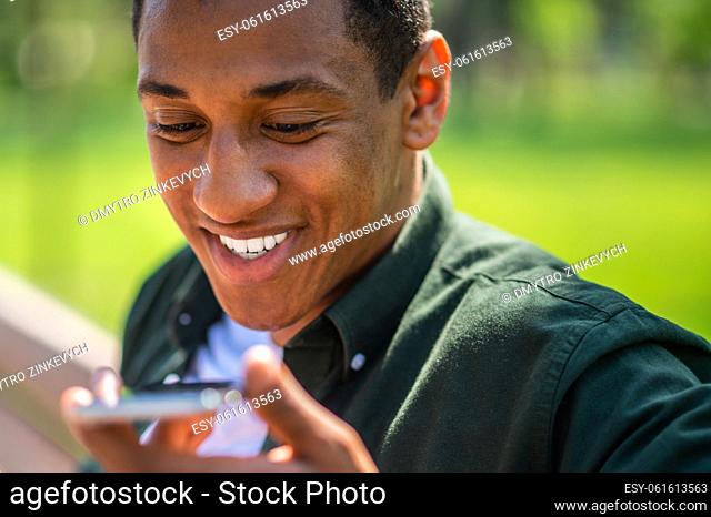 Positive emotions. Face of dark-skinned smiling young man saying message on smartphone outdoors on fine day