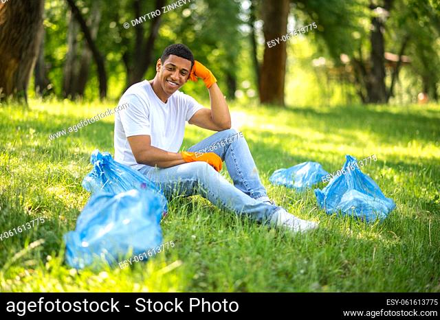Result of work. Dark-skinned guy in protective gloves sitting on grass smiling looking at garbage collected in bags in park