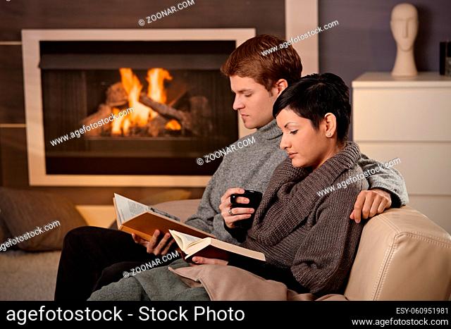 Young couple hugging on sofa in front of fireplace at home, reading books