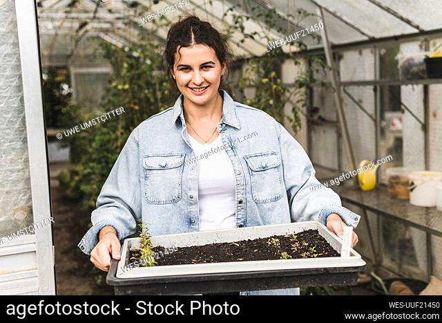 Smiling young woman holding tray while standing in greenhouse