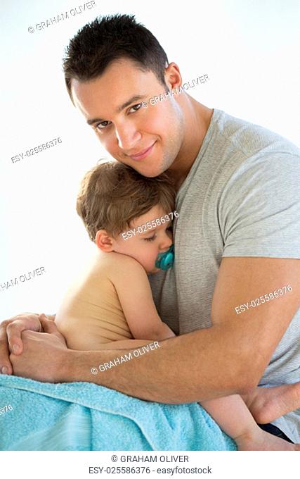 Sleepy baby boy cuddling his father, who is smiling at the camera with his arms wrapped round him