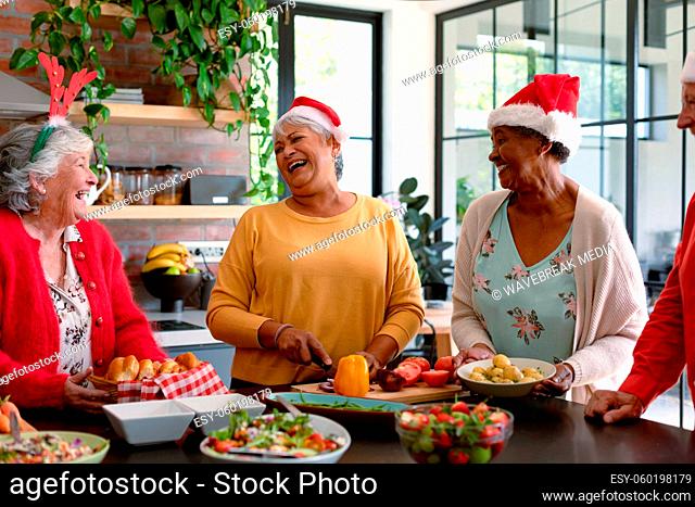 Four happy diverse senior male and female friends in christmas hats cooking together in kitchen