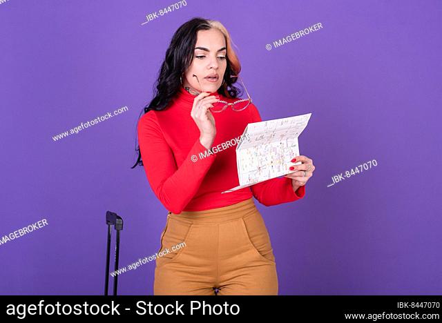 Young woman traveler with suitcase while reading a map against purple background. Studio shot