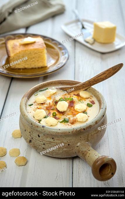 A bowl of homemade creamy potato and bacon soup topped with cheddar cheese, green onions, and oyster crackers with a side of cornbread