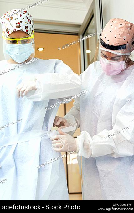 Doctor helping assistant wearing protective suit while standing at office