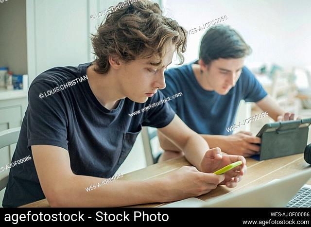 Two teenage boys using smartphone and tablet on table at home