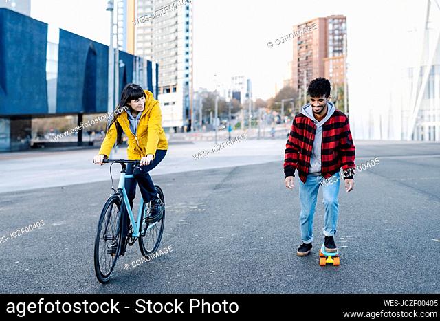 Smiling woman riding bicycle by friend skateboarding on road