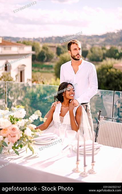 Mixed-race wedding couple. Destination fine-art wedding in Florence, Italy. African-American bride sits at wedding table, Caucasian groom hugs her shoulders