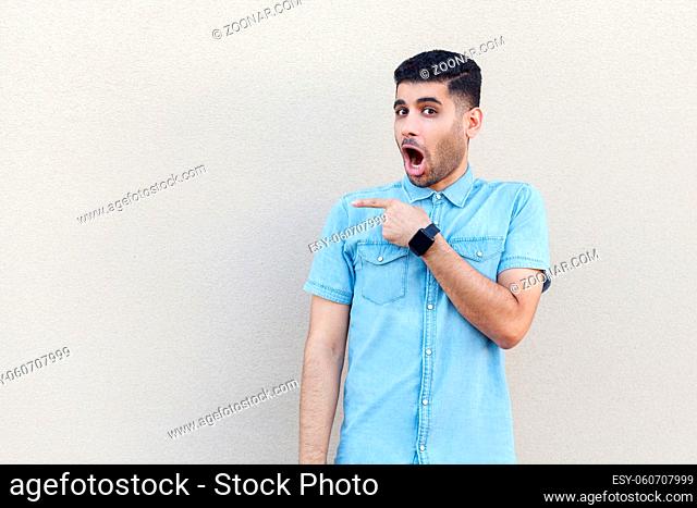 Portrait of shocked handsome young bearded man in blue shirt standing, looking at camera with surprised face and pointing at copyspace