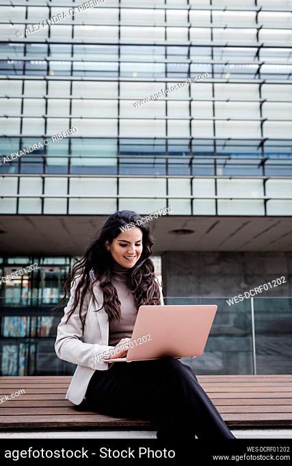 Smiling young businesswoman sitting on bench using laptop