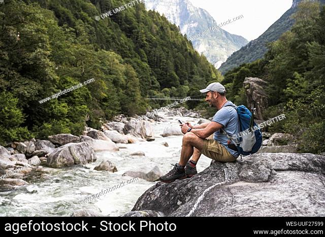Mature man using smart phone sitting on rock by river