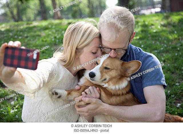 Young couple in park taking selfie whilst kissing corgi dog