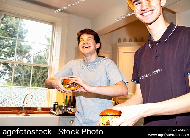 Portrait happy teenage boys eating hamburgers in kitchen