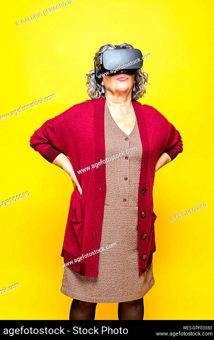 Senior woman with hand on hip wearing virtual reality headset while standing against yellow background