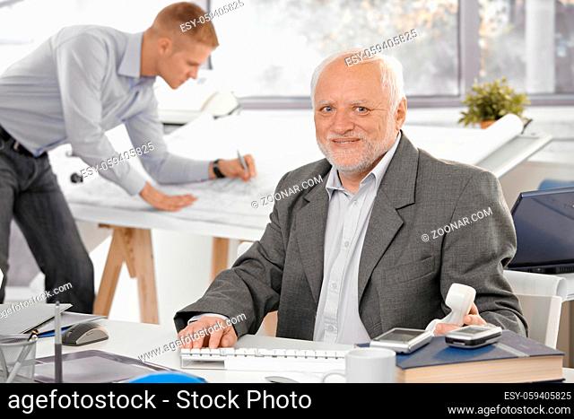 Senior businessman sitting at desk, smiling at camera, young architect working in office background
