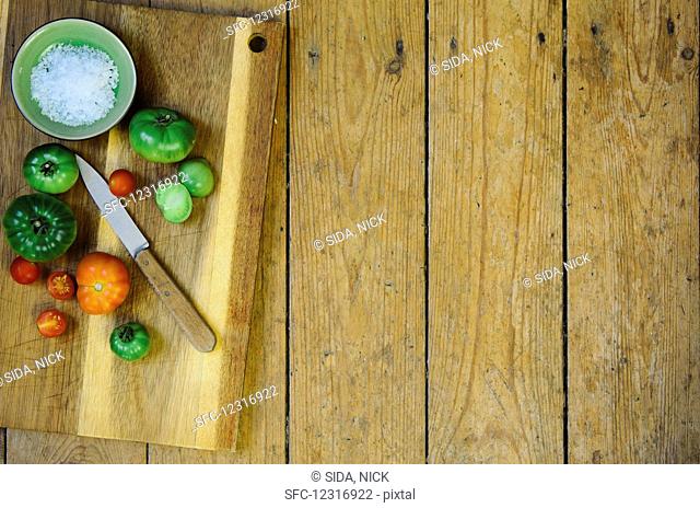 A bowl of salt and various tomatoes with a knife on a wooden chopping board