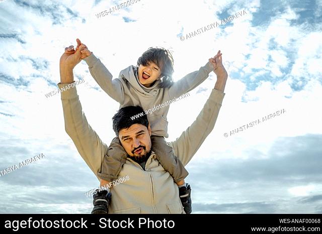 Happy son having fun sitting on father's shoulder in front of cloudy sky