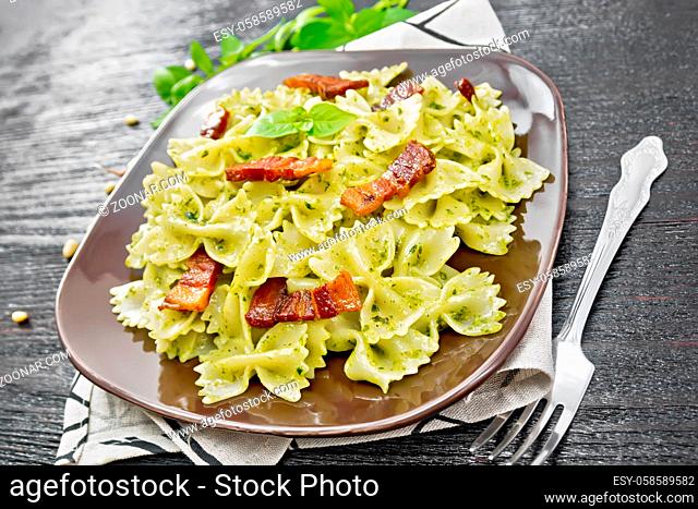 Farfalle pasta with pesto sauce, fried bacon and basil in a plate on a towel against dark wooden board