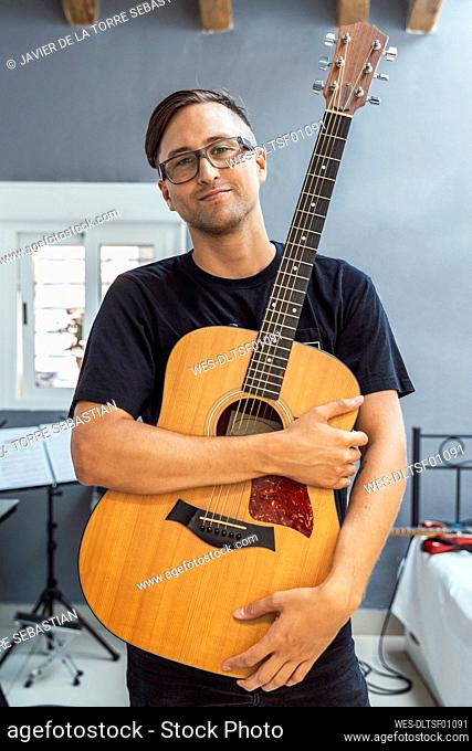 Confident musician holding guitar against wall at home