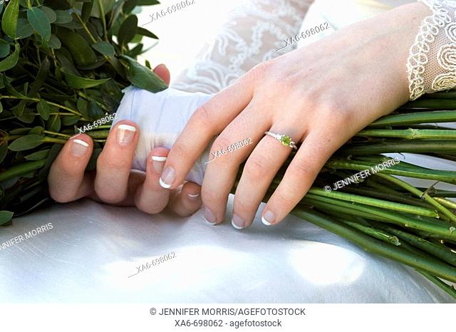 A close-up of a young bride's hands as she holds her wedding bouquet. Her lace sleeves are visible and she wears a ring with a green stone