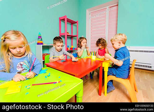 The cute preschoolers sit at the desks in the playroom and draw with colored pencils