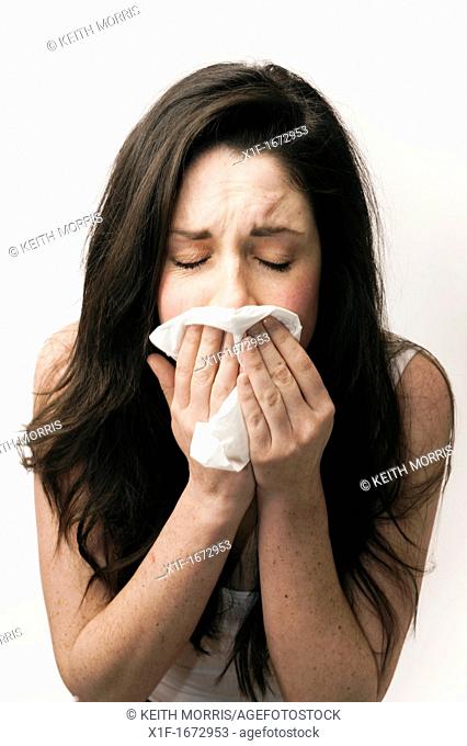 A young woman with brown hair and eyes feeling ill sneezing blowing her nose into a paper tissue