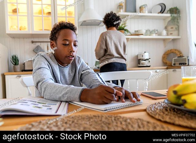 Boy writing on book with mother in background working at home