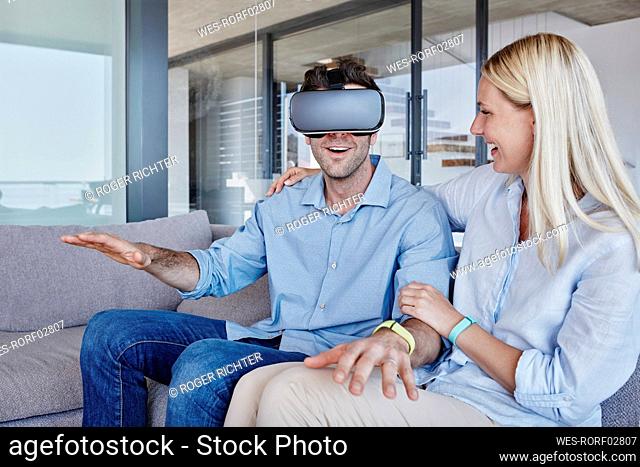 Cheerful woman looking at man wearing virtual reality simulator gesturing while sitting in living room