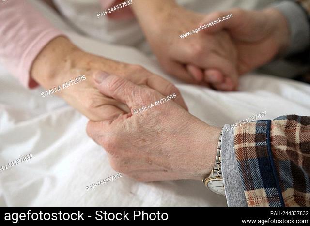 Aged man holding his wifes hands in bed on white bed sheet. Hands of old man with wrist watch holding old womans hands. || Model approval available