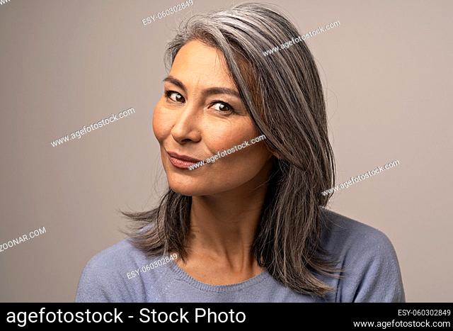 Gray Haired Mongolian Woman on a Gray Background. A Pensive Smile on a Woman's Face. Mongolian Beauty Concept. Close Up Shoot