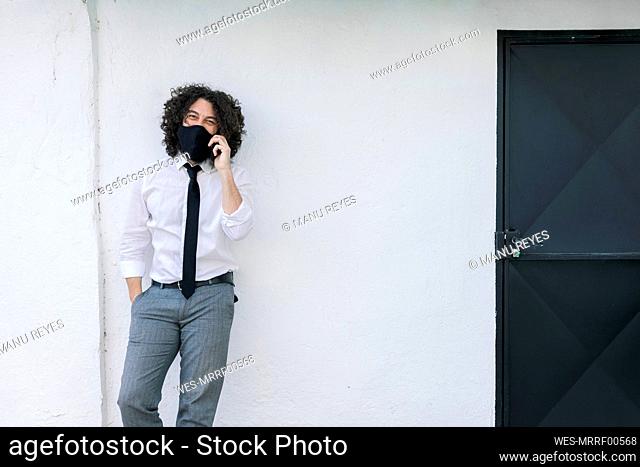 Businessman with face mask talking on mobile phone while standing against wall