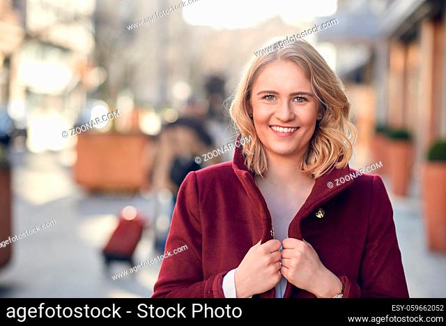 Attractive young blond woman with a friendly smile wearing a maroon overcoat walking a street with sunlight