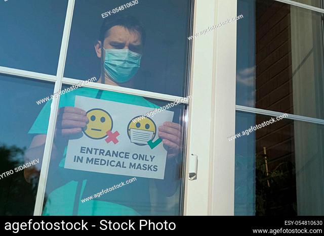 Announcement in a shop window: Entrance only in a medical mask. It is forbidden to enter without a medical mask. Store employee puts on a mask and attaches the...