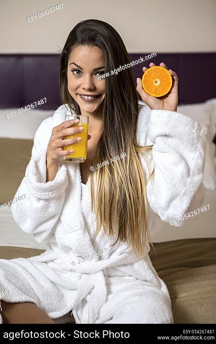 Pretty woman in white morning gown sitting on bed in room of hotel with glass of juice and cut orange and looking at camera