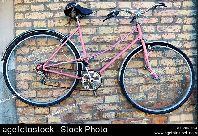 Old rusty broken pink women's bike hanging on a yellow brick wall