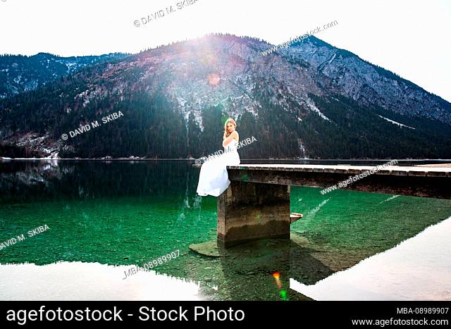 Young blond woman in bridal gown on a footbridge at the lake with mountains in the background, daylight