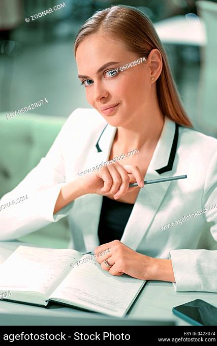 Beautiful businesswoman in white jacket looking at camera making notes in her scheduler or journal sitting in front at the modern bright office