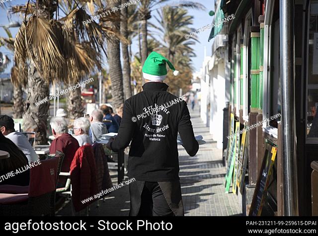 11 December 2023, Spain, Palma: A waiter at the ""Zur Krone"" restaurant on Arenal beach wears a Santa hat during a sunny day with a record temperature of 24...