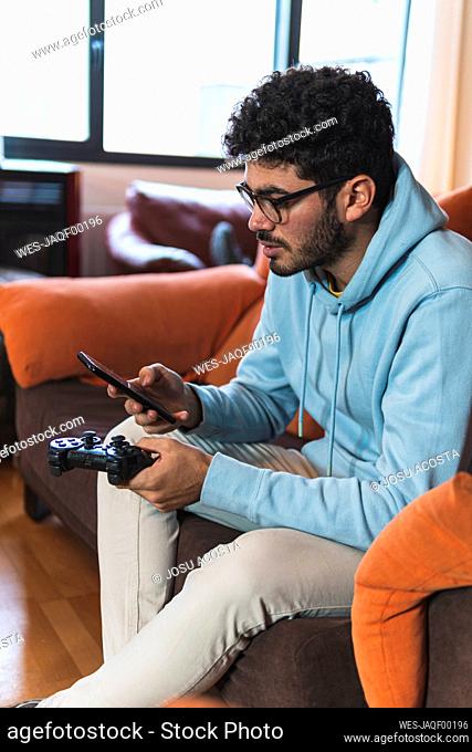 Young man using smart phone with game controller while sitting on sofa in living room