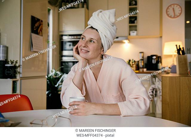 Portrait of smiling woman wearing a towel turban sitting with cup of coffee at table in the kitchen