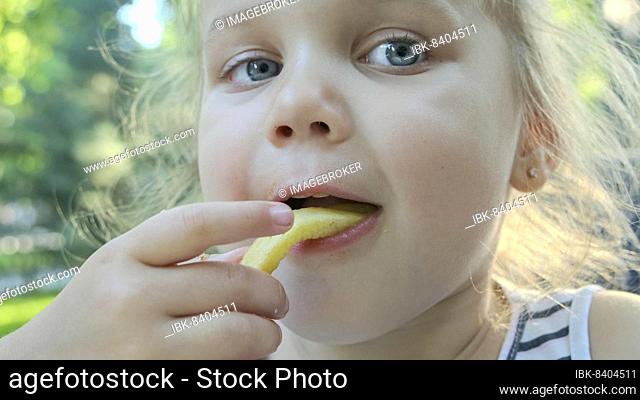 Little girl eat french fries. Close-up of blonde girl takes potato chips with her hands and tries them sitting in street cafe on the park