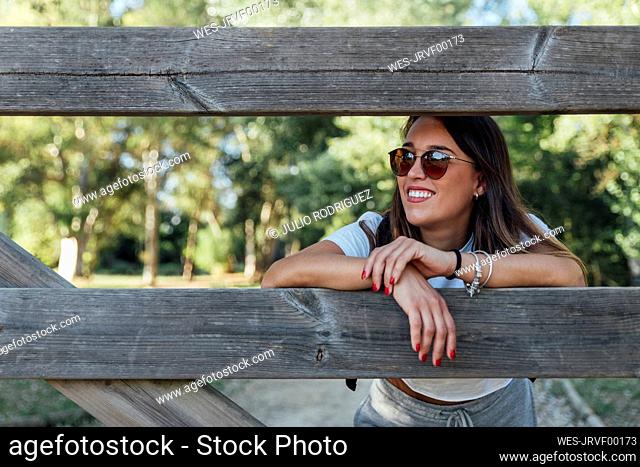 Smiling young woman wearing sunglasses leaning on wooden fence in forest