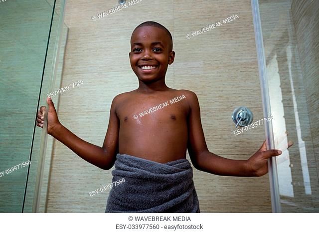 Low angle portrait of smiling shirtless boy wrapped in gray towel standing amidst glass