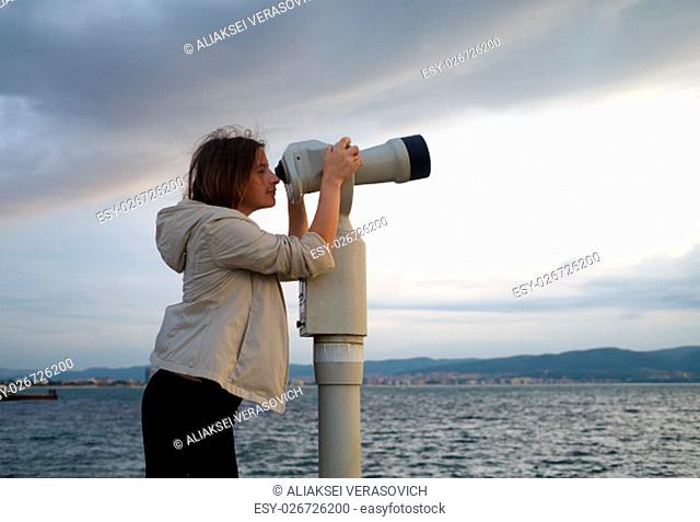 Pretty young woman using a coin operated binocular enjoying a great view of the Black sea. Early morning. Shallow depth of field. Focus on the model