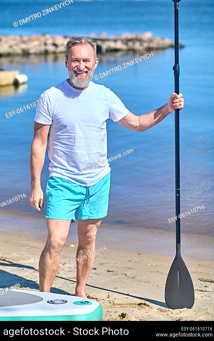 Best time. Happy bearded middle aged man with paddle looking confidently at camera standing barefoot on sand near water