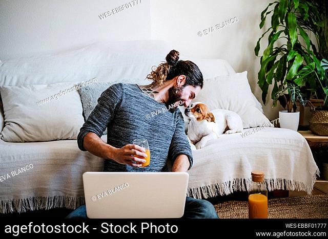 Man with juice and laptop smiling while looking at dog sitting on sofa at home