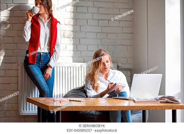 Beautiful young blonde haired woman making some notes in her textbook sitting in front of the laptop her frend drinking coffee on the background copyspace cafe...