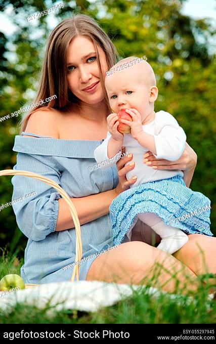 young happy mom with her baby on hands sitting at grass in park, smiling, baby eats apple
