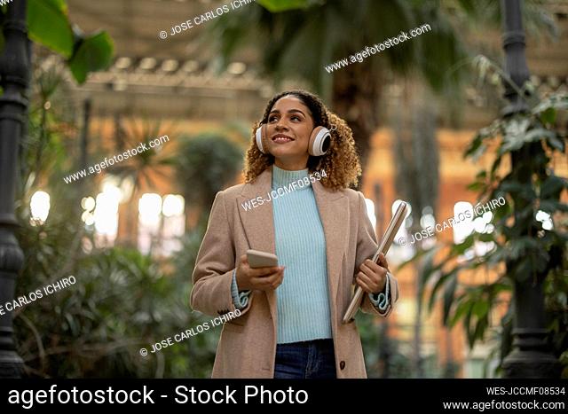 Happy businesswoman listening to music through wireless headphones holding laptop and mobile phone